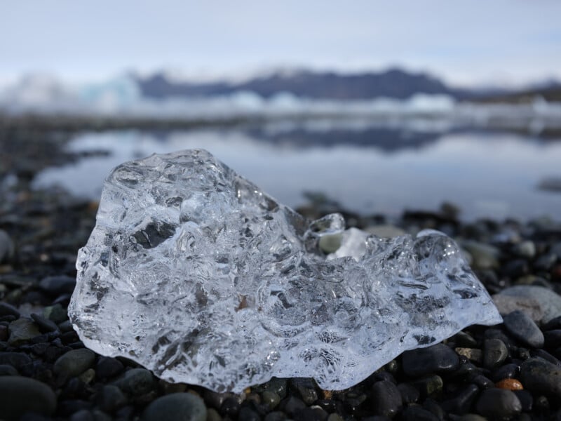 A clear chunk of ice rests on a rocky shoreline, with a blurred background of water and distant mountains under a cloudy sky.