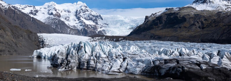 A large glacier with jagged ice formations stretches into a murky glacial lake, surrounded by steep, rocky mountains and snow-covered peaks under a partly cloudy sky.