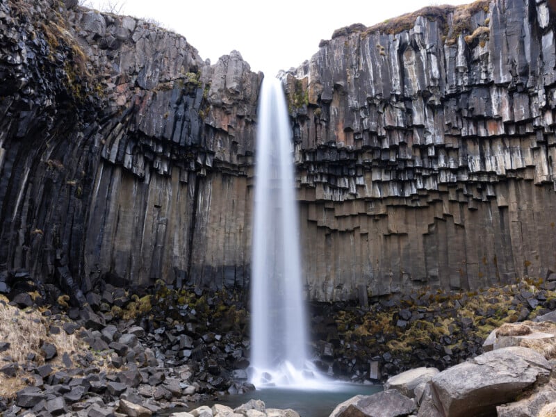 A tall, narrow waterfall flows over a cliff of striking hexagonal basalt columns, surrounded by rocky terrain and sparse vegetation under an overcast sky.