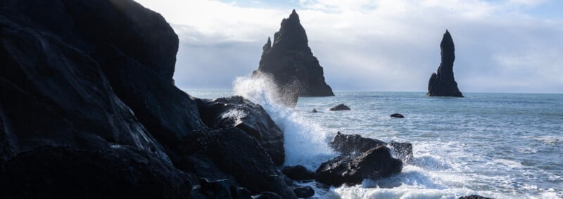 Waves crash against dark, jagged rocks on a coastline, with two towering sea stacks rising from the ocean under a cloudy sky.