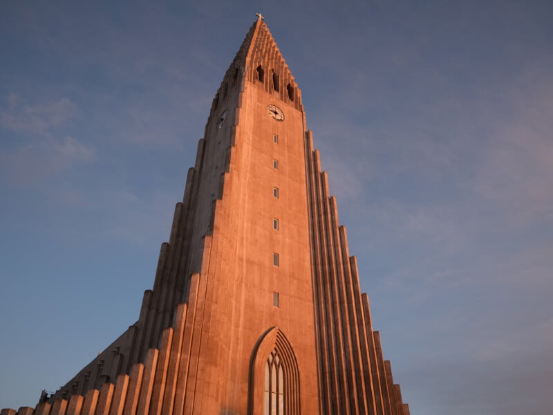 A tall, modern concrete church with a stepped, symmetrical facade and a central clock tower, lit by warm sunlight against a partly cloudy sky.