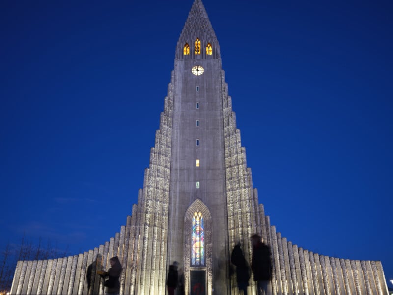 A tall, illuminated church with a pointed, tiered façade and a clock near the top stands against a deep blue sky at dusk. Blurred silhouettes of people are seen walking in front of the entrance.