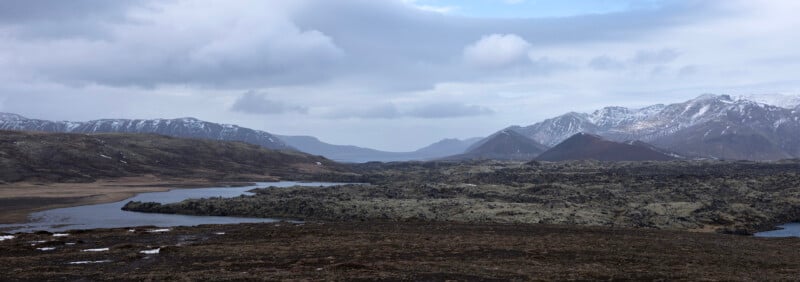 A wide, rocky landscape with patches of snow on distant mountains under a cloudy sky, featuring a winding river or lake cutting through the rough terrain.