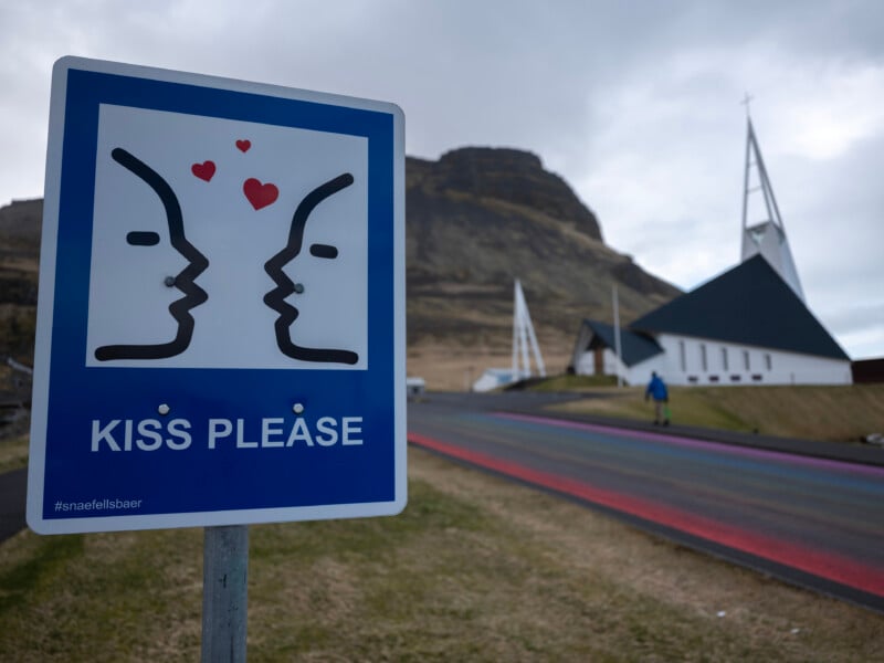 A blue sign with two cartoon faces about to kiss and hearts above them says “KISS PLEASE" near a road, with a church and mountain in the background.