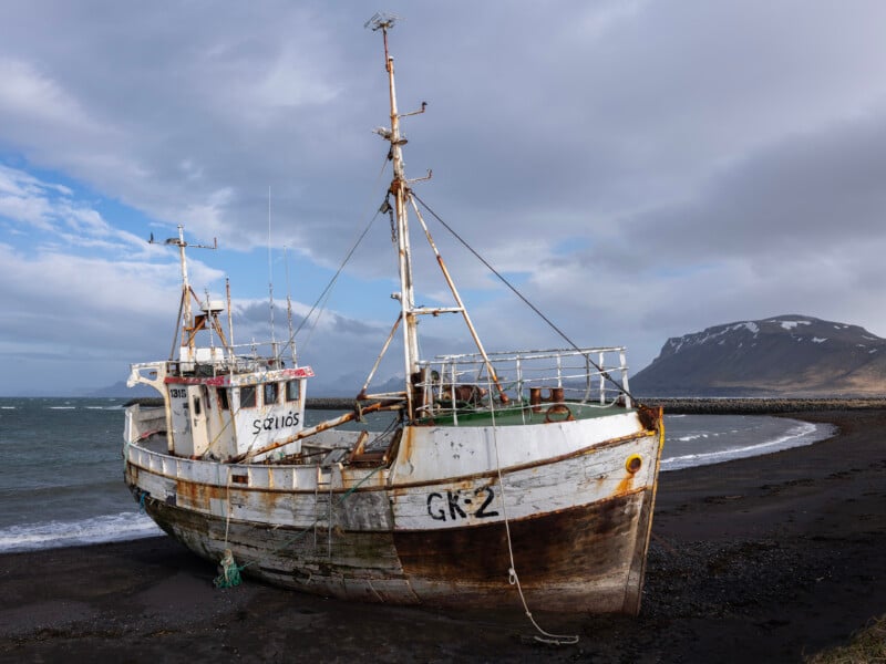 An old, rusted fishing boat labeled "GK2" rests abandoned on a black sand beach, with mountains and cloudy skies in the background.