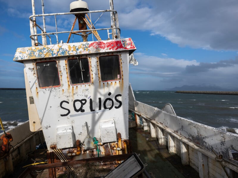 A rusty, abandoned fishing boat with the word “SOLIÓS” painted on its cabin sits tilted on a shore, with choppy sea and distant mountains visible under a partly cloudy sky.