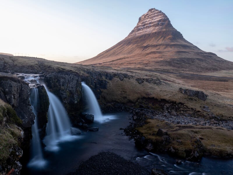 A tall, conical mountain rises behind a small waterfall cascading into a rocky stream, surrounded by brown grasslands under a clear sky.