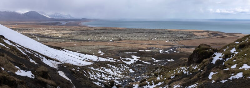 Panoramic view of a coastal landscape with patches of snow on rocky hills, a winding road, flat brown fields, and distant mountains under a cloudy sky by the sea.
