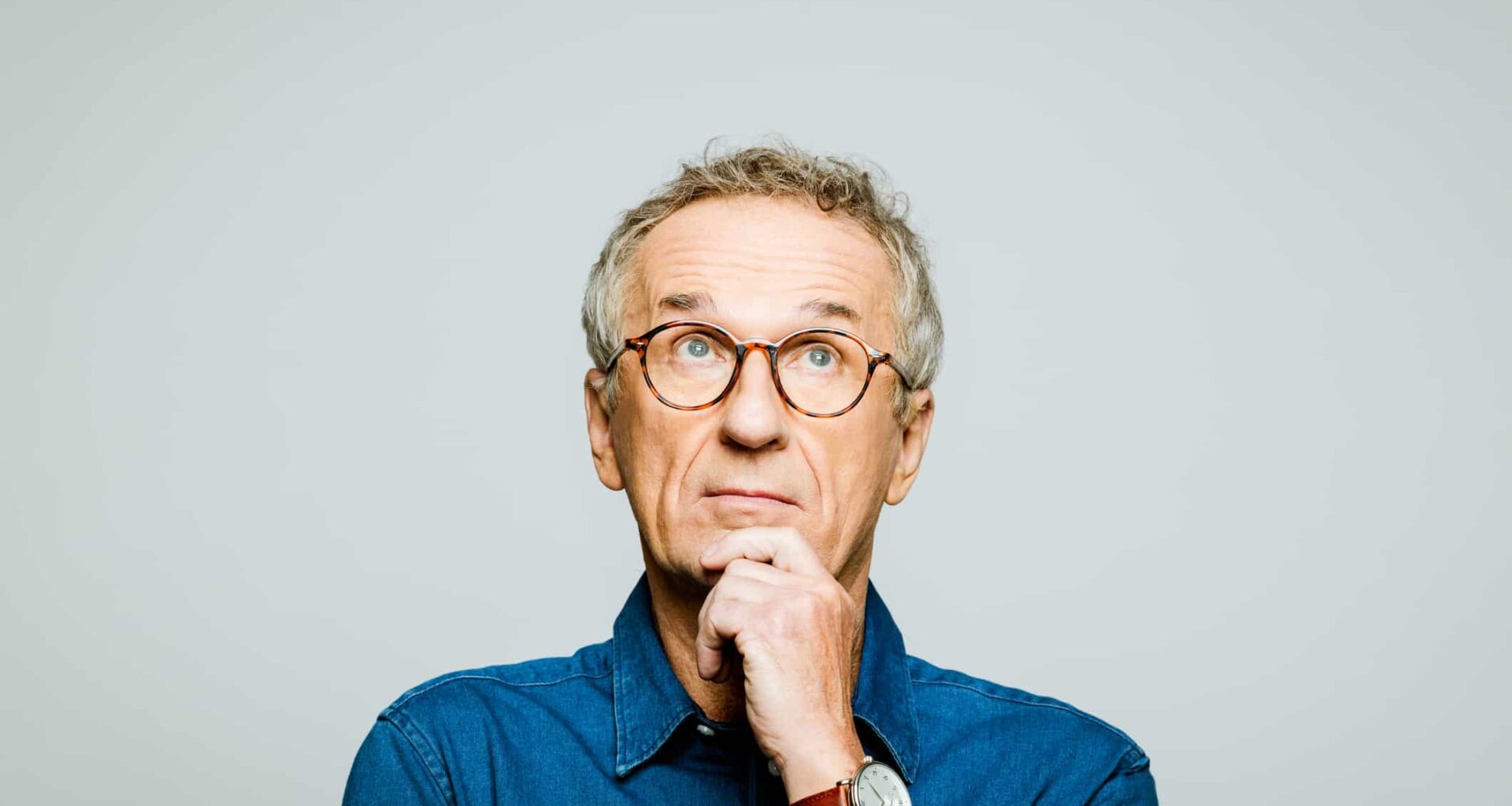 Portrait of elderly man wearing white denim shirt and glasses looking up with hand on chin. Thoughtful senior entrepreneur, studio shot against grey background.