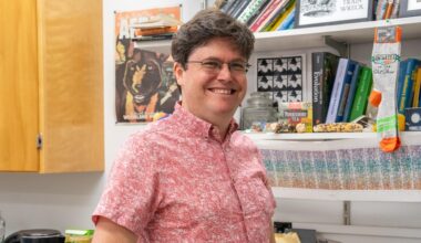 Head and upper body of a white man with short dark hair and glasses wearing a pink short sleeved shirt and smiling. Behind him are bookshelves.