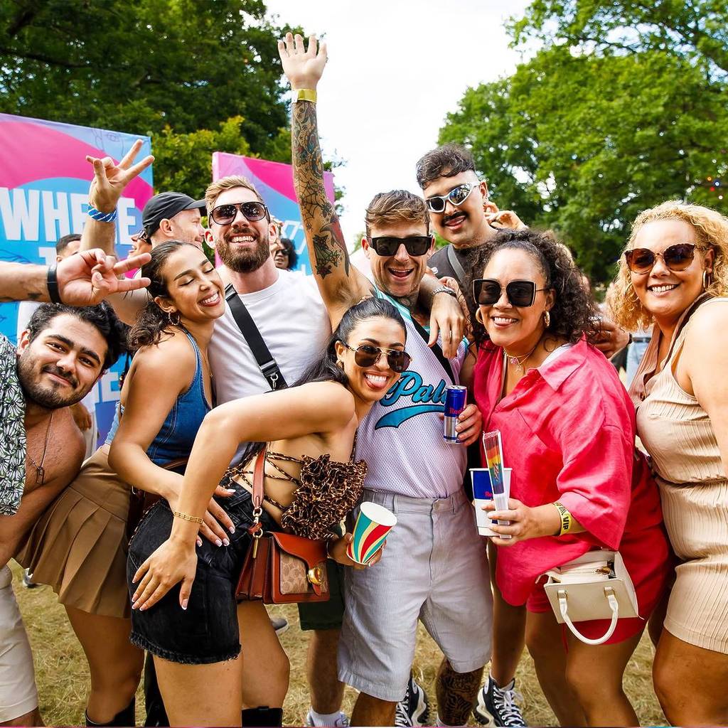 A crowd of friends smile, holding drinks up in a rowdy summer festival celebrating LGBT culture.