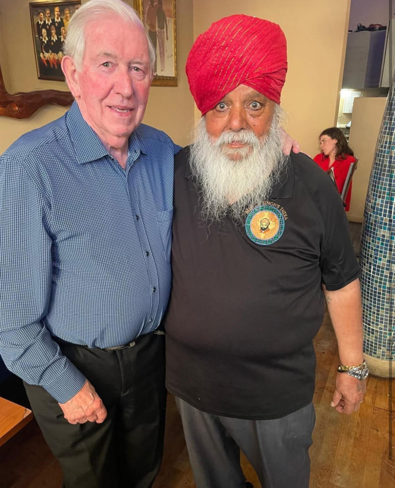 Two men posing for a photo; one man is wearing a blue shirt and the other is wearing a red turban and black shirt.