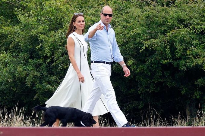 A man and woman walk a black dog on a path, pointing towards something, surrounded by dense green foliage.