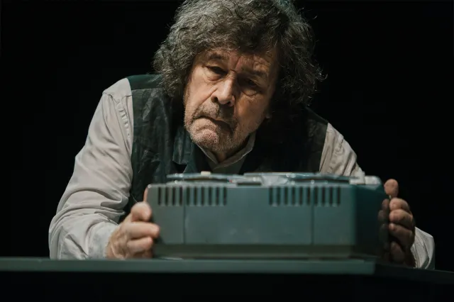 An actor sits on stage behind a tape recorder.