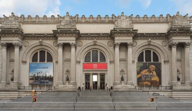The grand entrance of the Metropolitan Museum of Art in New York City, featuring wide stone steps, arched doorways, columns, and large banners advertising exhibitions on either side of the main entrance.