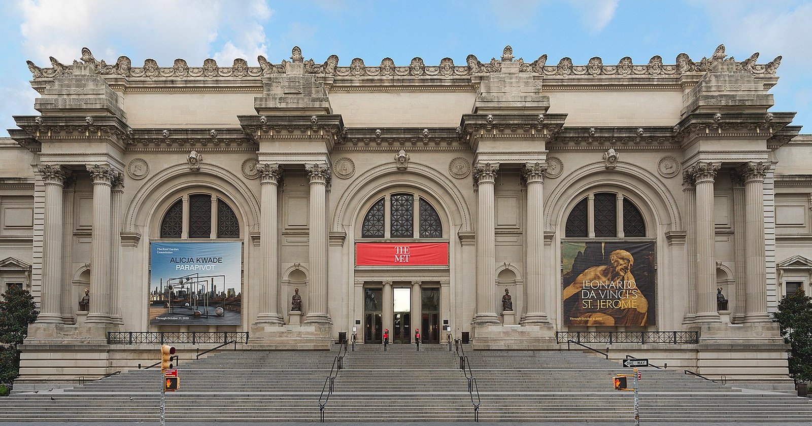 The grand entrance of the Metropolitan Museum of Art in New York City, featuring wide stone steps, arched doorways, columns, and large banners advertising exhibitions on either side of the main entrance.
