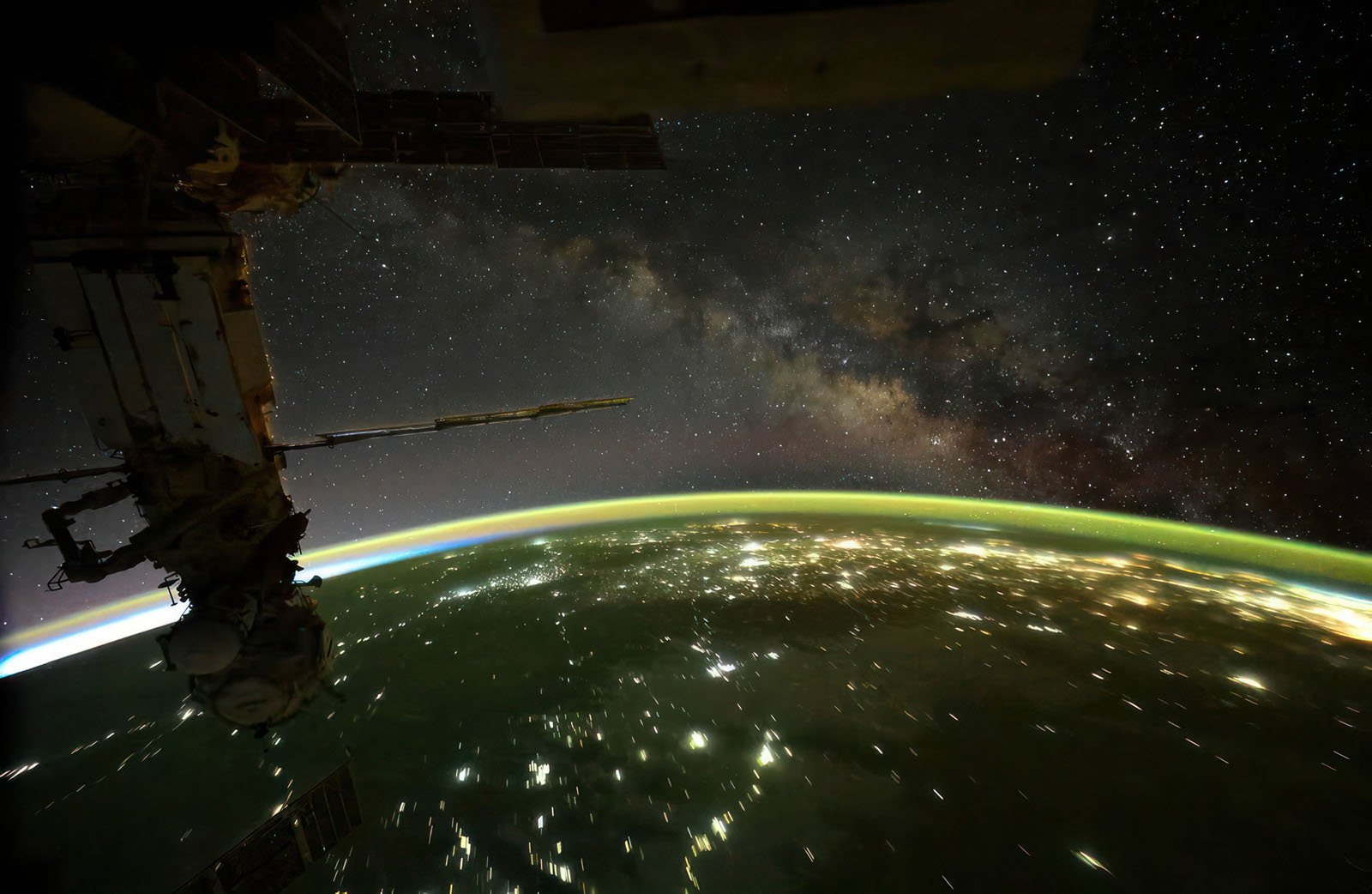 View from space showing part of a space station, city lights on Earth's surface, a glowing green atmospheric layer, and the Milky Way galaxy stretching across the star-filled sky.