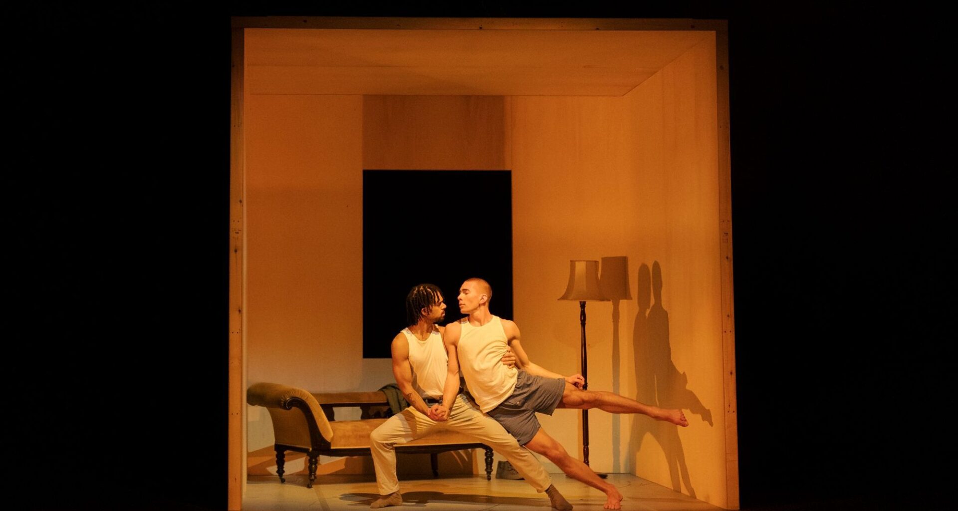 Two male dancers in an intimate pose, the weight of one man's body holding up the other. They stand within a small cube representing Giovanni’s room. The room also contains a chaise lounge and a lampstand. The photo is a scene from the dance theatre production 'Inside Giovanni’s Room'.