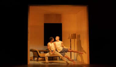 Two male dancers in an intimate pose, the weight of one man's body holding up the other. They stand within a small cube representing Giovanni’s room. The room also contains a chaise lounge and a lampstand. The photo is a scene from the dance theatre production 'Inside Giovanni’s Room'.