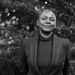 A black-and-white photograph of a Black woman in a vest standing amid trees.