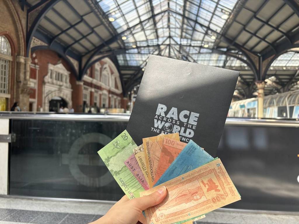 a hand holds up a Race Across the World envelope with a fan out banknotes inside Liverpool Street Station