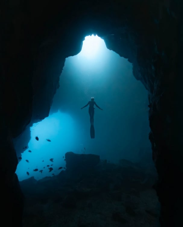 A diver wearing fins floats upward toward the light at the entrance of an underwater cave, surrounded by rocks and a few fish, with a blue glow illuminating the scene.