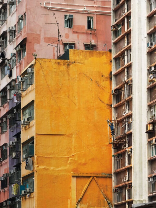Tall apartment buildings with small windows and balconies, some painted in bright yellow, pink, and orange hues. Many windows have air conditioning units, and laundry is hanging outside. A man is working on scaffolding.
