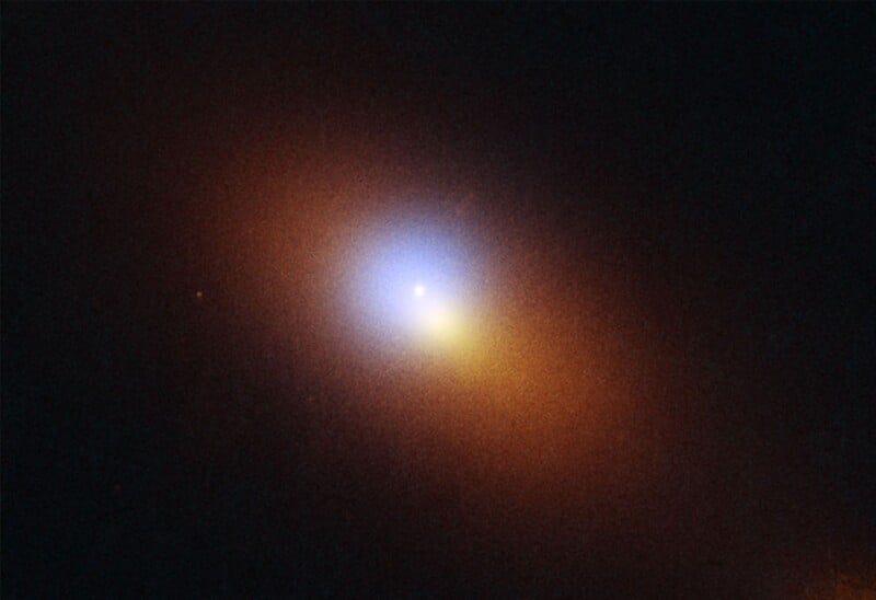 A bright, glowing spot with a blue-white center fades outward into orange and red hues, surrounded by a dark background, resembling a galaxy or nebula in space.