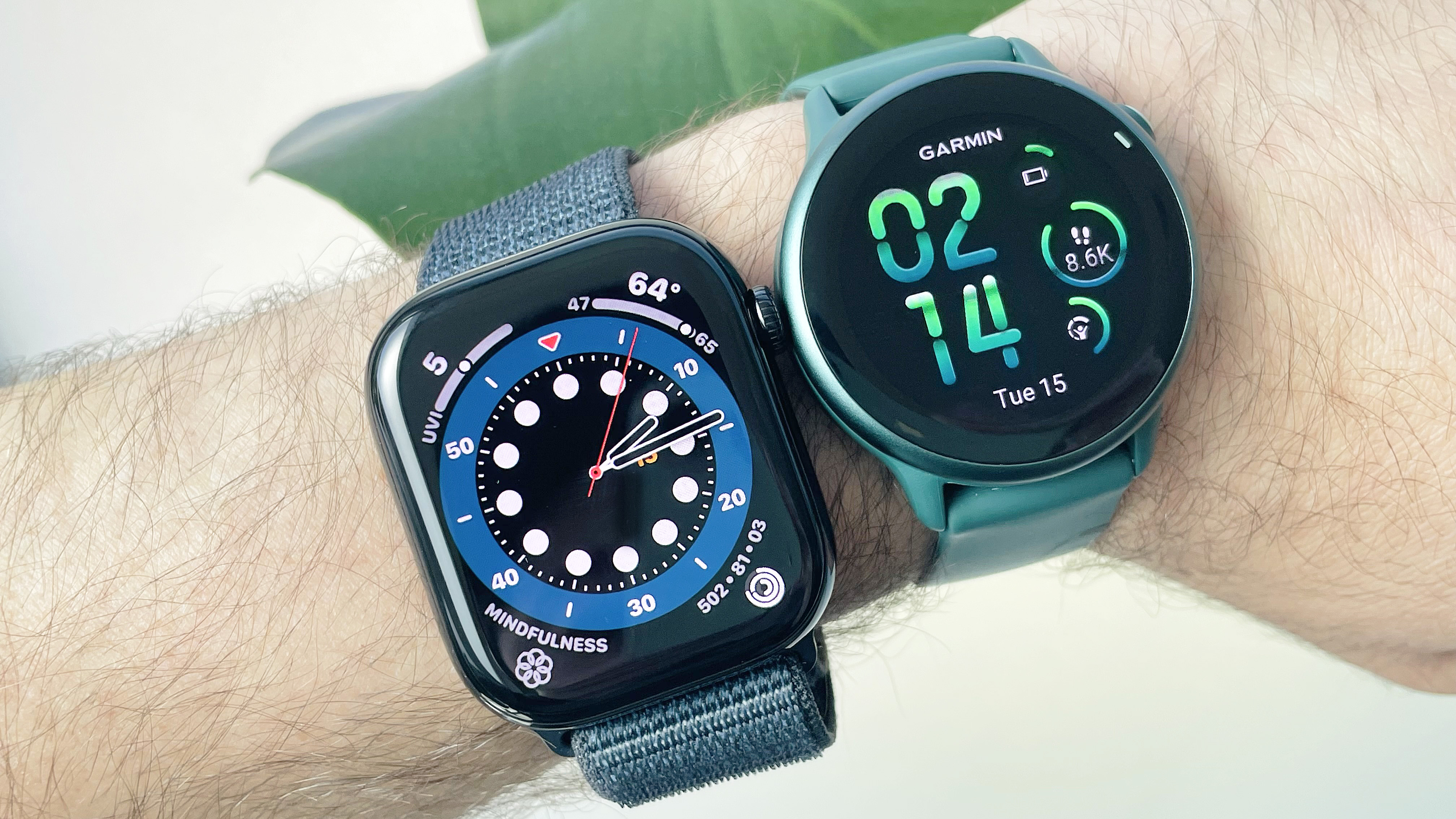 Close-up of a user's wrist wearing both the Apple Watch 10 on a blue fabric strap (left) next to the Garmin Vivoactive 6 (right) on an emerald green strap