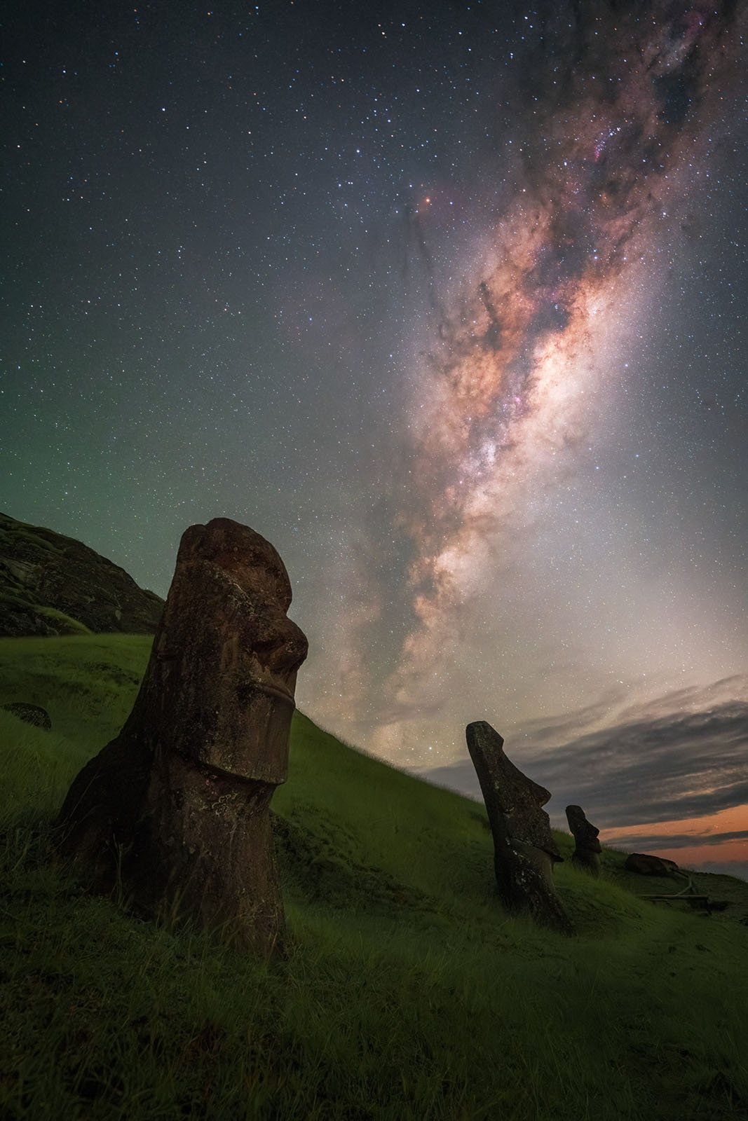 Moai statues on a grassy hill under a star-filled night sky, with the Milky Way galaxy brightly visible and stretching diagonally across the background.