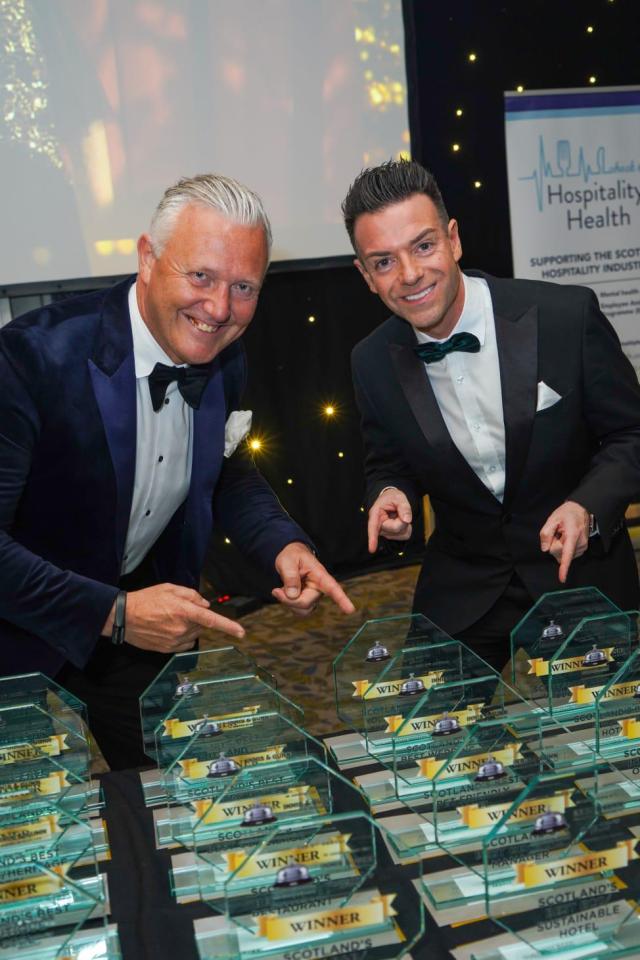 Two men in tuxedos pointing at a row of awards.