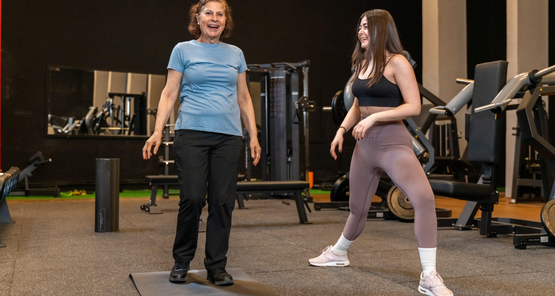 Two women in a gym. A younger woman demonstrates a leg position while the older woman looks forward smiling