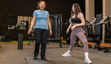 Two women in a gym. A younger woman demonstrates a leg position while the older woman looks forward smiling