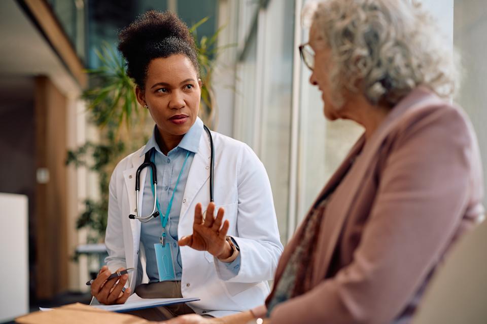 Doctor in a lab coat attentively listening and talking to an older woman in formal attire during a consultation in a bright office setting