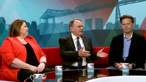 BBC Julie Minns, Lord Kirkhope, and Andrew Husband sitting on the BBC Politics North red sofa. Minns, with a brown bob cut and wearing an orangey red half sleeve jacket and black top looks to her left with her face contorted slightly as if she is mid laugh or cry. In the middle with receding but still mainly light brown hair, Kirkhope wears the standard dark suit, dark tie, light shirt combo. He's mid sentence and has both hands stretched out in front of him. His expression could be that of someone hoping to convince, but might be exasperation. Husband has gone office casual. Black jacket over a blue shirt with the top buttons undone. Grey trousered, no tie, neat brown hair that has a fair amount of volume. With his hands lightly clasped in front of him, his mouth is turned up at one side, perhaps showing a lack of agreement with Kirkhope, or perhaps just confusion. There are four BBC Politics branded mugs on the table in front of them, perhaps for thirst avoidance, perhaps for aesthetically repetitive marketing. 