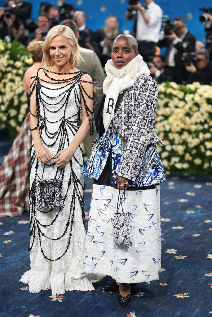 Tory Burch, wearing a white floor-length lace gown with black beading draped overtop, poses with Janice Bravo, wearing a black, white and blue suit jacket, with an embroidered neckpiece and a white sequined skirt.