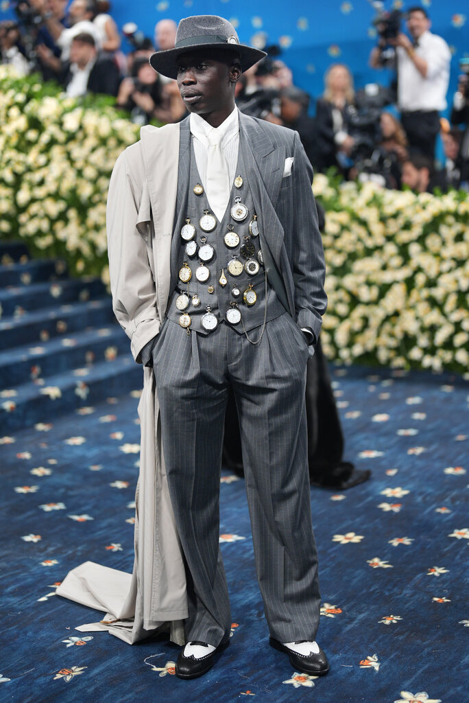 Khaby Lame wearing a grey pinstripe suit with pocket watches attached to the waistcoat, a beige trench coat draped over one shoulder, a grey wide-brim felt hat, and black and white wingtip shoes. 