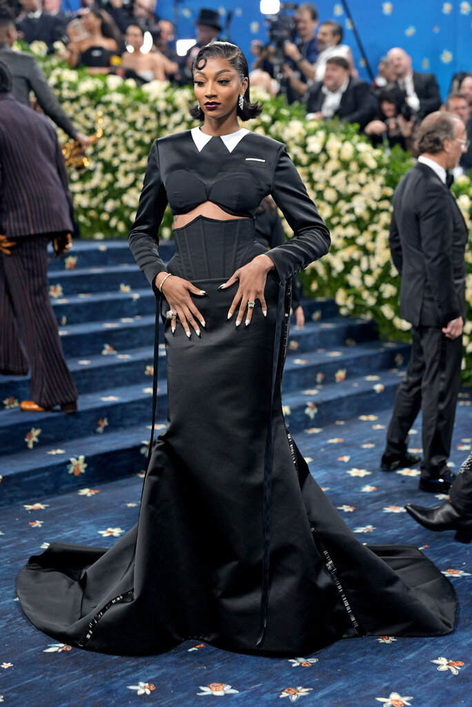 Angel Reese wearing a black blouse, cropped just below the bust line, with a white collar and long-sleeves, paired with a corseted floor-length black skirt.