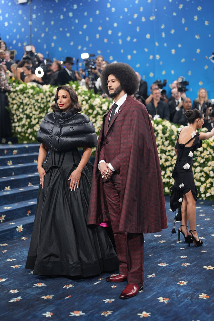Colin Kaepernick is wearing a maroon plaid suit with a matching cape and Nessa Diab is wearing a black dress with a puffer capelet.