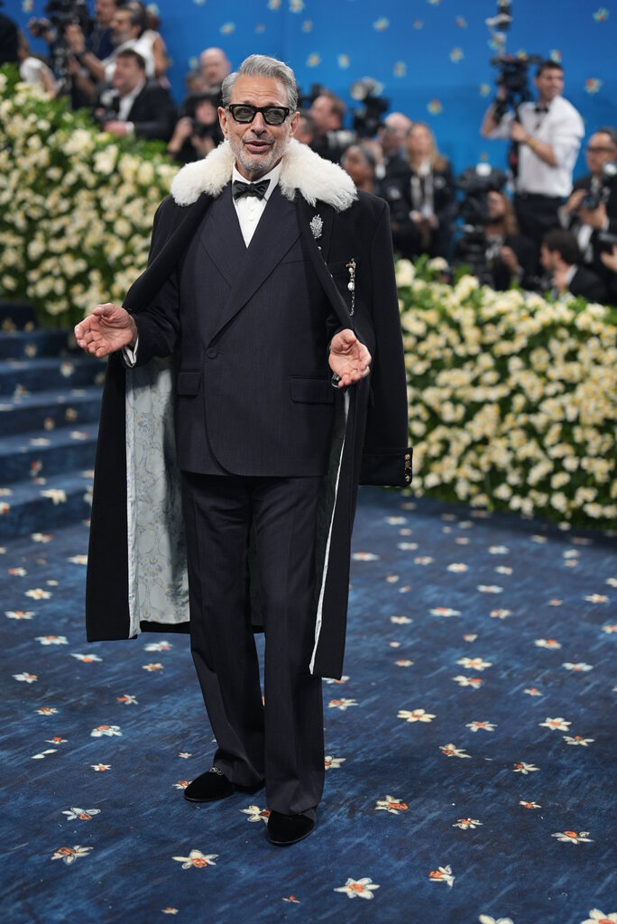 Jeff Goldblum wearing a black suit, with a bowtie and an overcoat embellished with brooches on the label and a white fur-like collar.