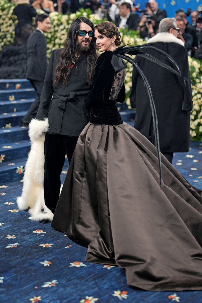 Alessandro Michele, wearing a black suit and tinted sunglasses, and Lana Del Rey, wearing a long brown silk skirt and feathered velvet top.