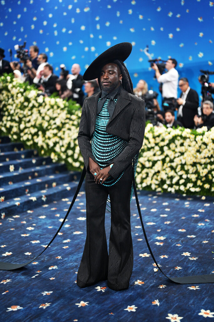 Shaboozey wearing a black suit with a cropped jacket, overtop a draped top made from blue beads, and a wide brim hat pinned at an angle.