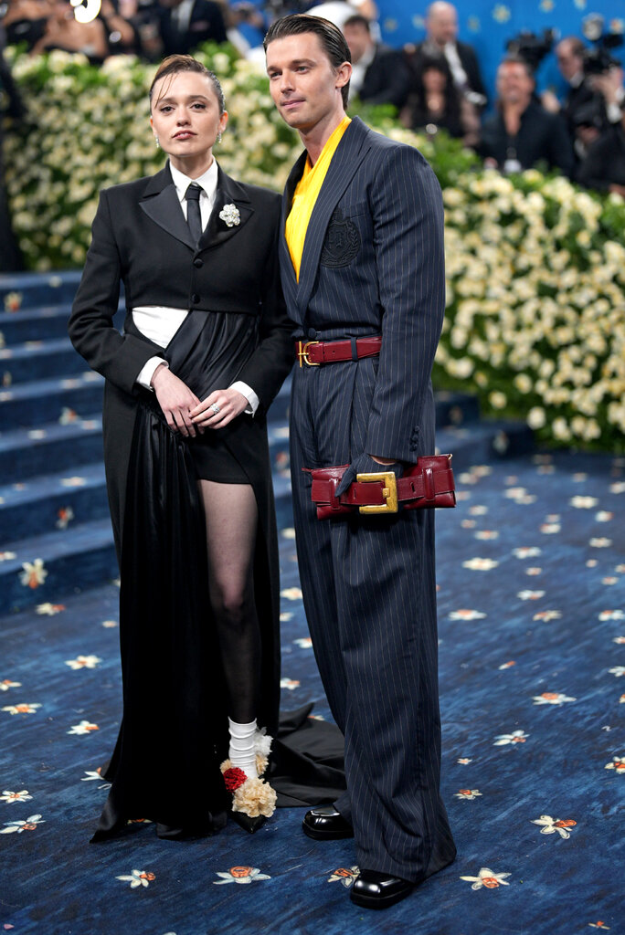 Aimee Lou Wood is wearing a black cropped suit jacket and with draped black details and Patrick Schwarzenegger is wearing a blue pinstripe suit and yellow shirt.