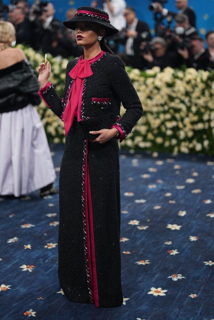 Whitney Peak, wearing a black tweed collarless jacket with hot pink detailing overtop a coordinating floor-length skirt, and matching hat, poses holding a black velvet clutch in one hand and what appears to be a cigarette in the other.