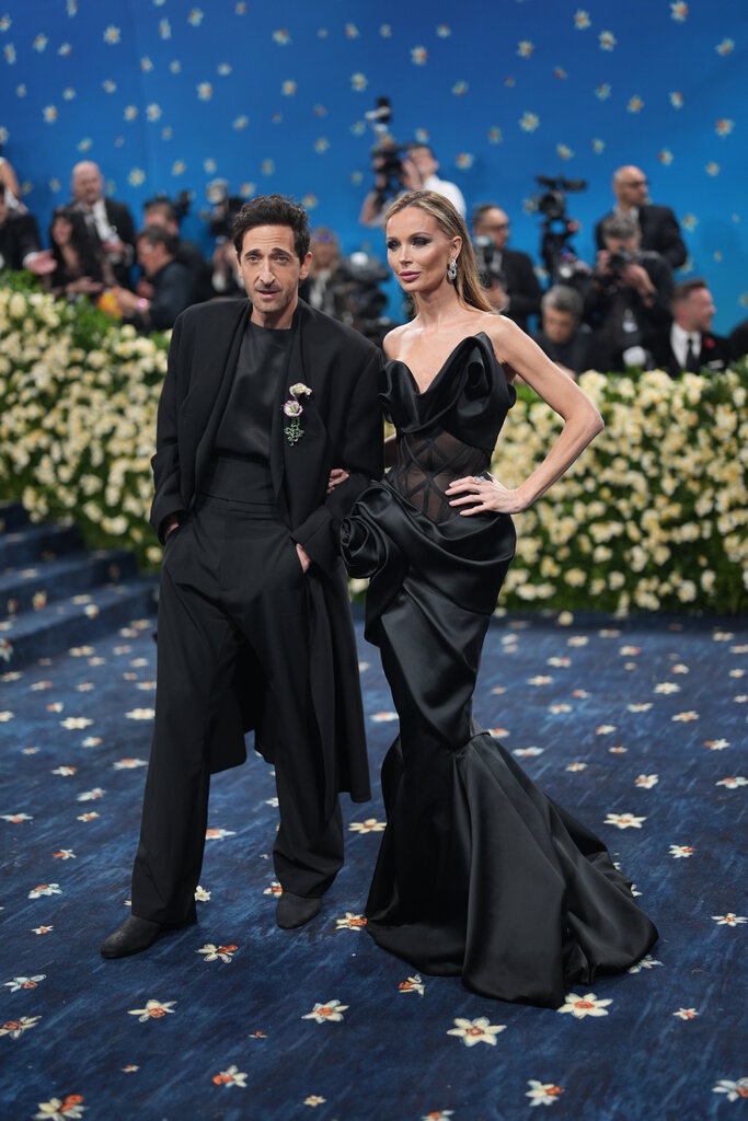 Adrien Brody, wearing a black draped overcoat and collarless top with trousers, poses with Georgina Chapman, wearing a sleeveless black mermaid gown, with an exposed corset and rosette detail at the hip.