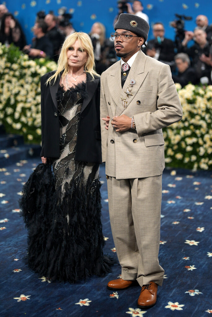 Donatella Versace, wearing a floor-length beige and black dress with feather detailing and a black suit jacket, poses with Chance the Rapper, wearing a beige double-breasted tuxedo with jeweled brooches on the lapel and a grey brimless felt hat.