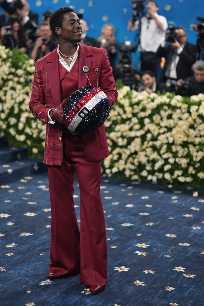 Damson Idris is wearing a red suit with matching leather gloves and holding a jeweled helmet.