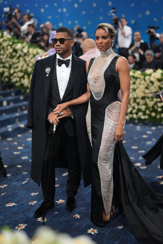Russell Wilson is wearing black tuxedo with a coat draped over his shoulders and Ciara is wearing a black dress with chains draped up the sides and front.