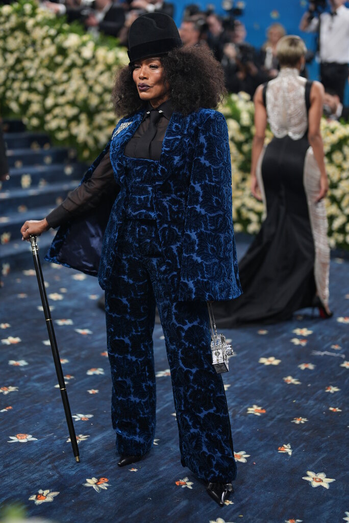 Angela Bassett is wearing a blue patterned suit and black hat and posing with a cane.