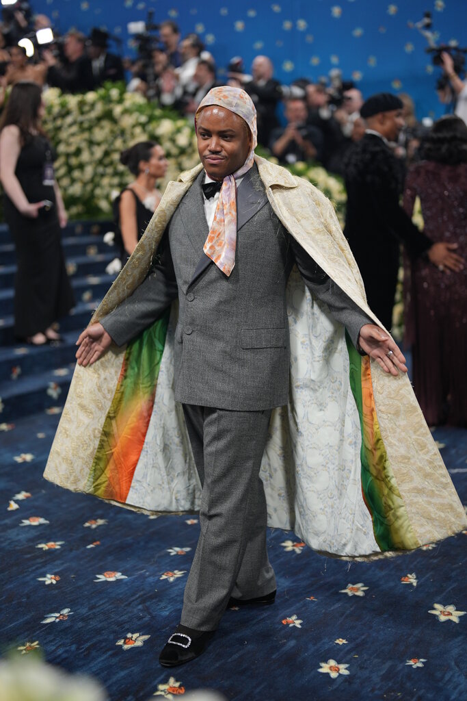 Eric Mack wearing a gray suit, a long yellow jacket and a light pink scarf around his head.